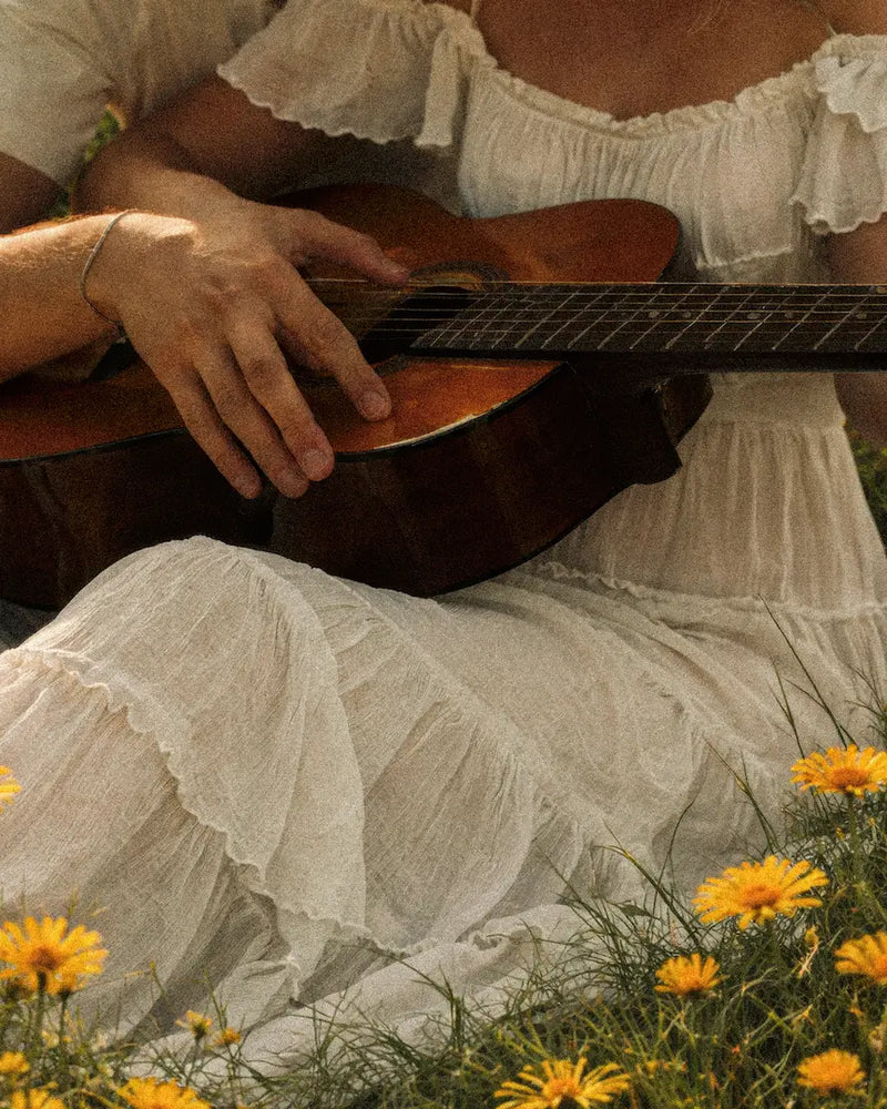 Person playing an acoustic guitar with a white dress and yellow flowers in the foreground